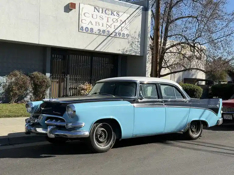 A light blue and black 1950s Dodge Custom Royal car is parked in front of a building with a faded sign that reads 'NICKS…