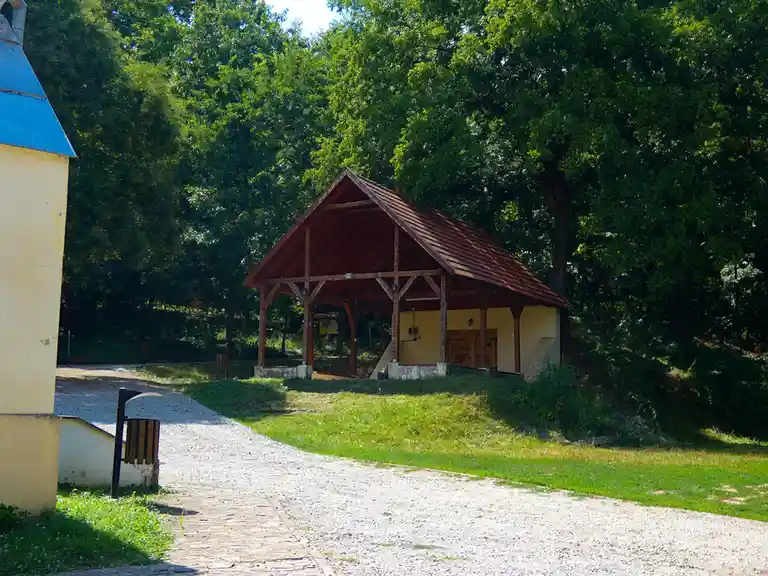 A gravel path leads to a wooden-roofed structure with yellow walls and wooden doors, surrounded by green trees and grass…