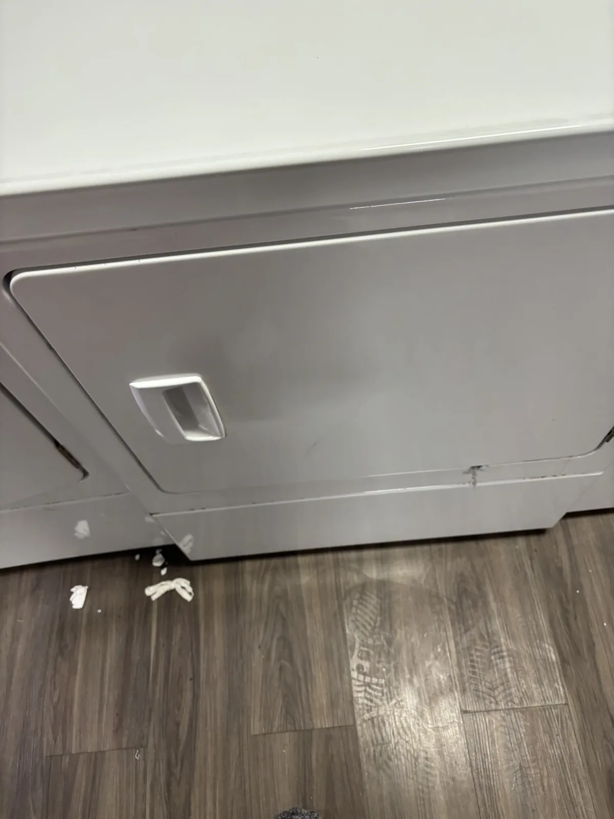 A close-up photo shows the bottom of a white front-loading dryer sitting on a dark wood-patterned floor, with small white…