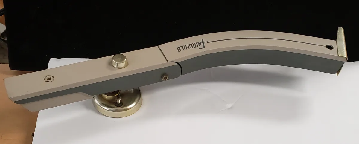 A close-up of a beige and gray Fairchild brand monitor arm, mounted on a brass base and attached to a black monitor, with…