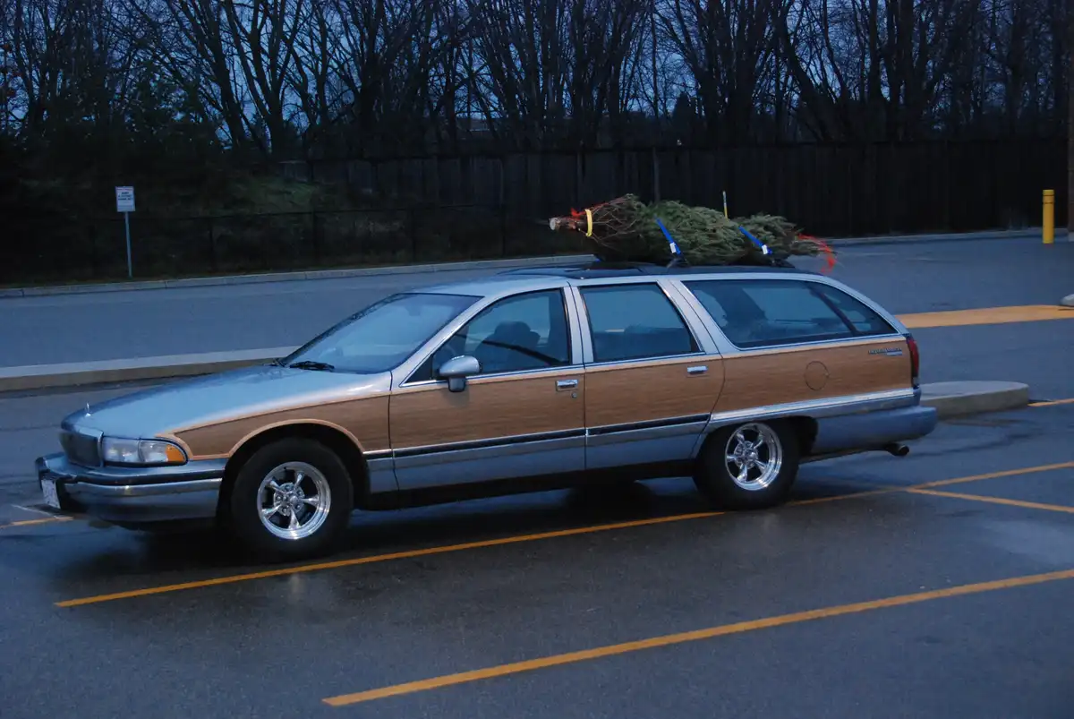 A silver and wood-paneled station wagon is parked in a lot with a large, untrimmed Christmas tree strapped to its roof rack…