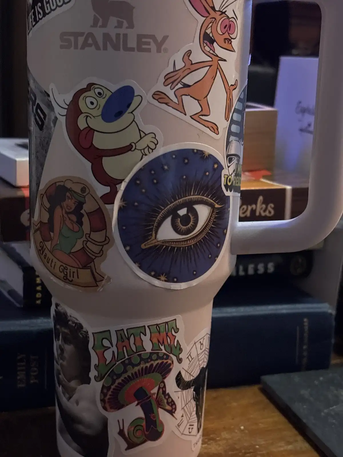 A white Stanley insulated mug is heavily covered in various colorful stickers, including cartoon characters like a brown…
