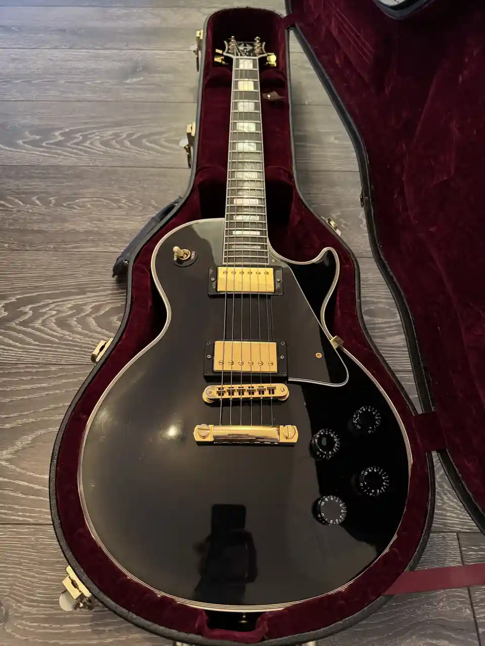 A black electric guitar with gold hardware rests inside an open, burgundy-lined hard case on a gray wood floor. The guitar…