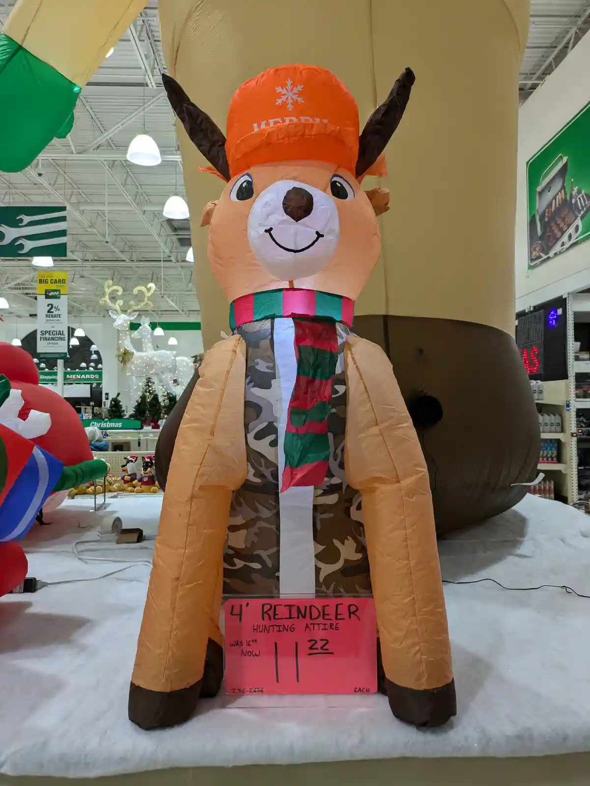 An inflatable reindeer decoration in a retail store, wearing a camouflage vest, a red and green striped scarf, and an orange…