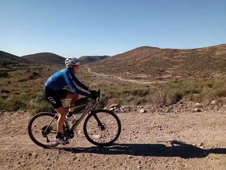 A cyclist in a blue and black cycling outfit with a white helmet rides a gravel bike on a dusty, rocky trail through a dry…