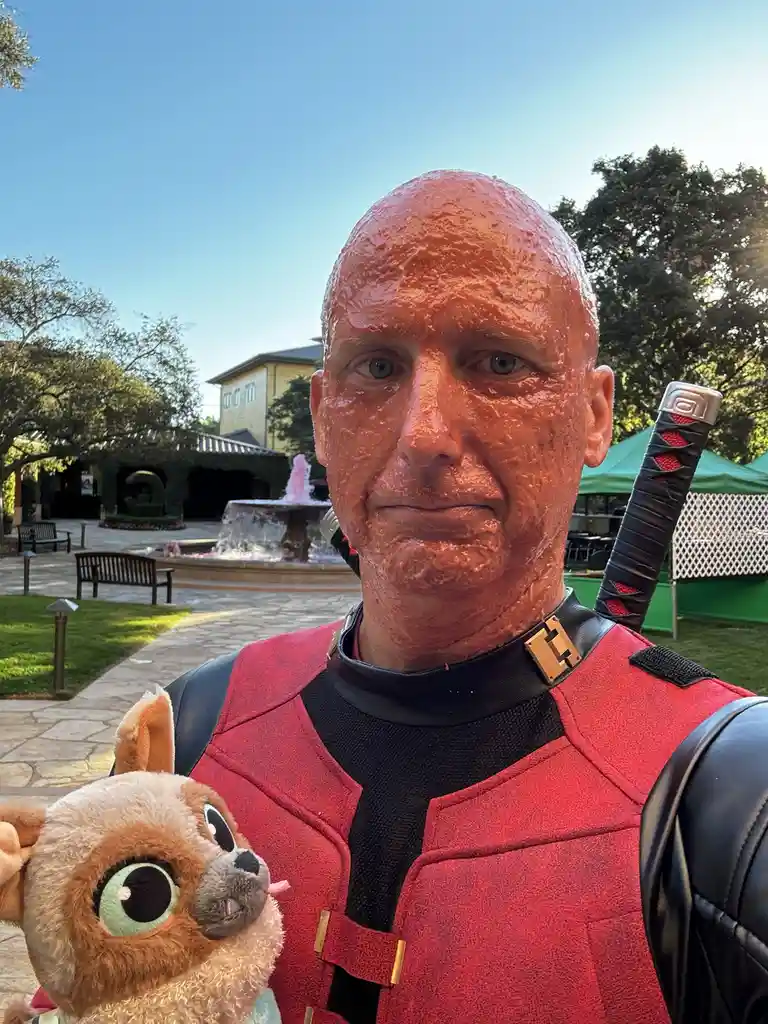 A man dressed in a red and black Deadpool costume with a katana on his back stands outdoors, his entire face covered in a…