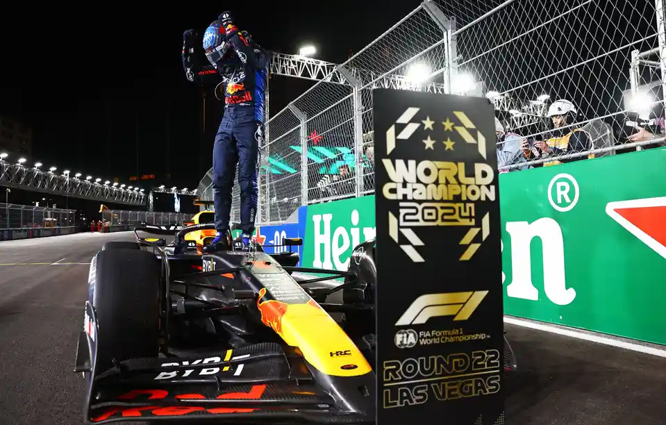 A Formula 1 driver in a Red Bull racing suit stands triumphantly on top of his black and yellow race car at night, arms…