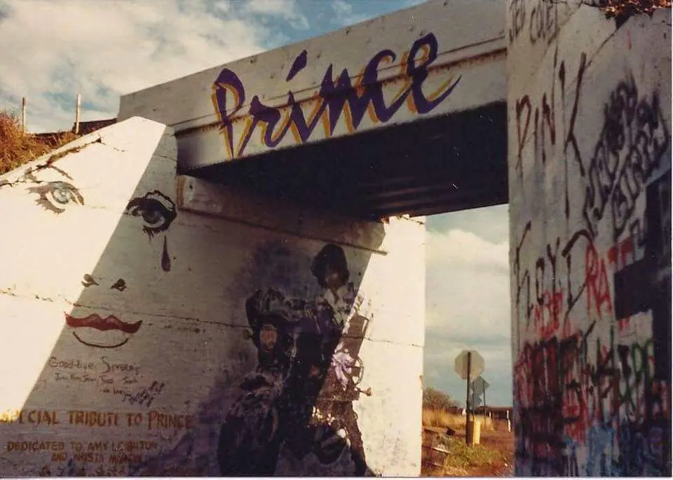 A weathered concrete bridge or overpass covered in graffiti, with the name 'Prince' painted in large purple cursive across…