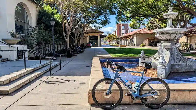 A light blue Cervélo road bike is parked on a low stone ledge next to a dry, ornate fountain in a sunny, tree-lined plaza…
