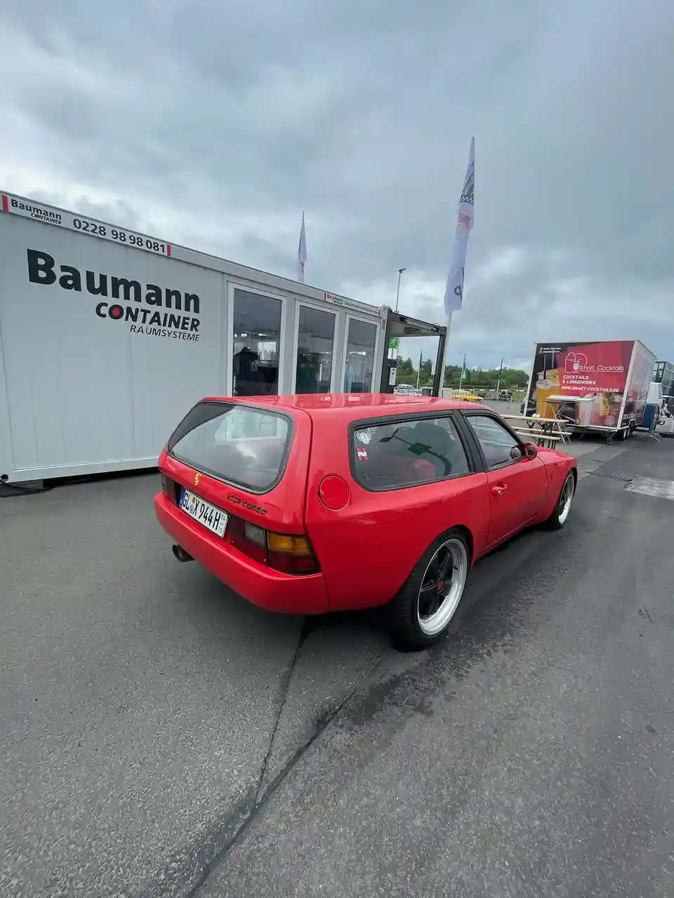 a red ferrari 208 gts with a station wagon body style is parked on asphalt next to a white 'Baumann Container' building…