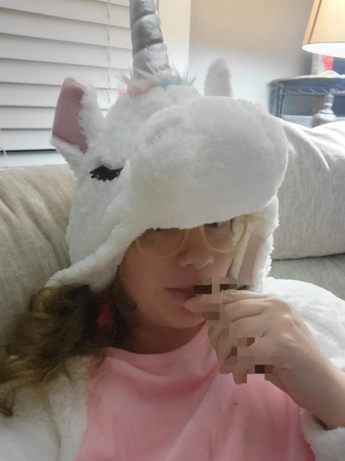 A person wearing a large, fluffy white unicorn hat with a horn and ears sits on a couch, holding a blurred object to their…