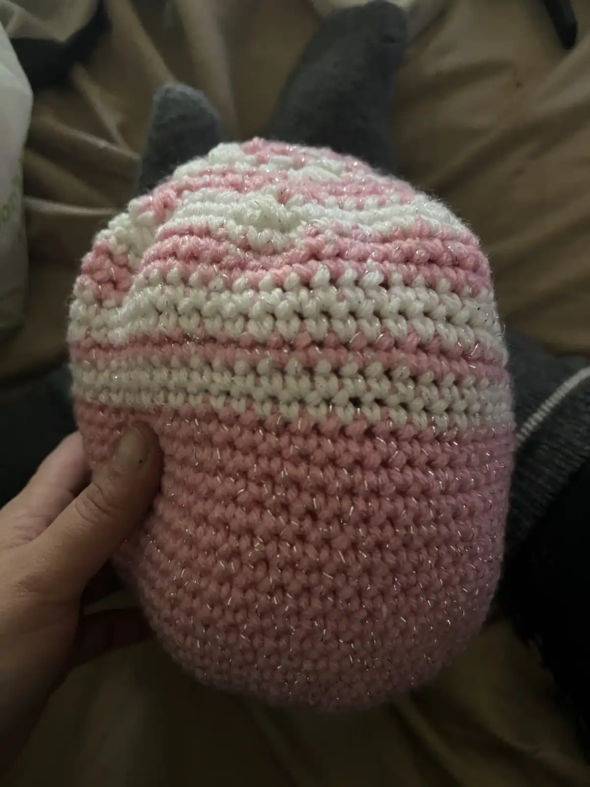 A hand holds a pink and white crocheted hat with sparkly yarn, featuring two dark gray cat ears on top. The hat is being…