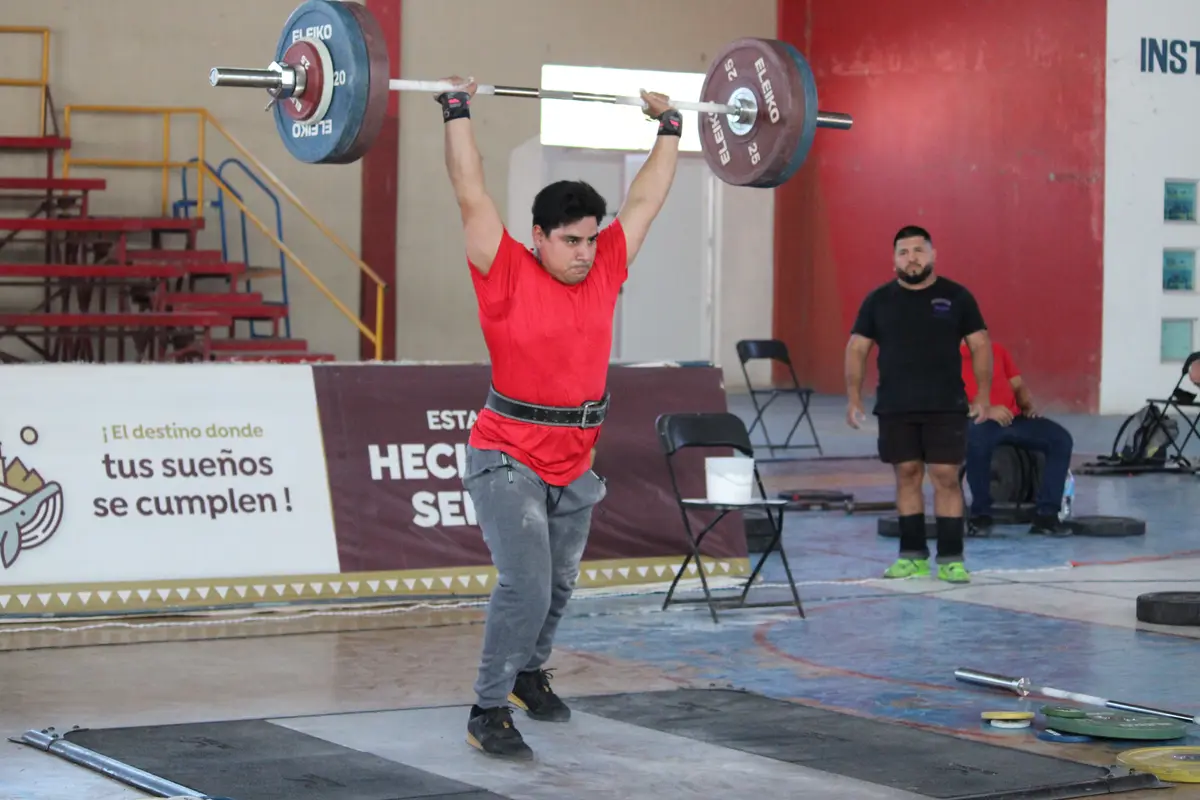 A male weightlifter in a red shirt and grey pants holds a barbell overhead in a gym, with a banner behind him reading '¡El…