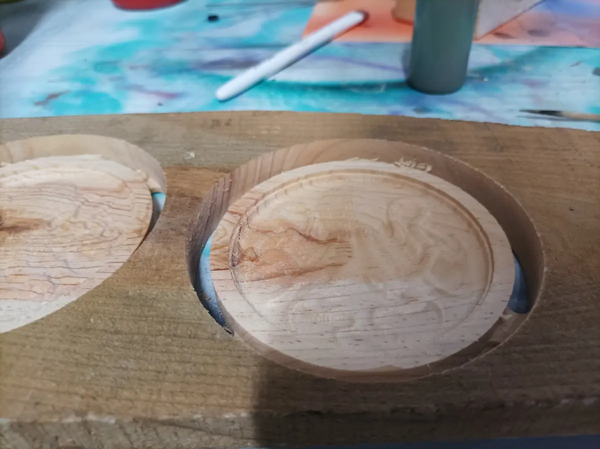 A close-up photo shows two circular wooden bowls carved into a larger wooden block, with visible wood grain and carving…