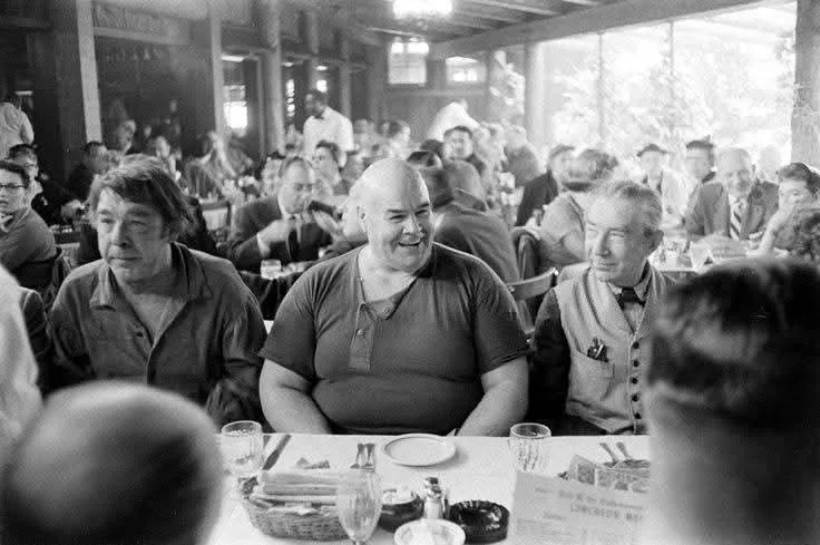 A black-and-white photo shows three men seated at a restaurant table, with the central, bald man grinning broadly while the…