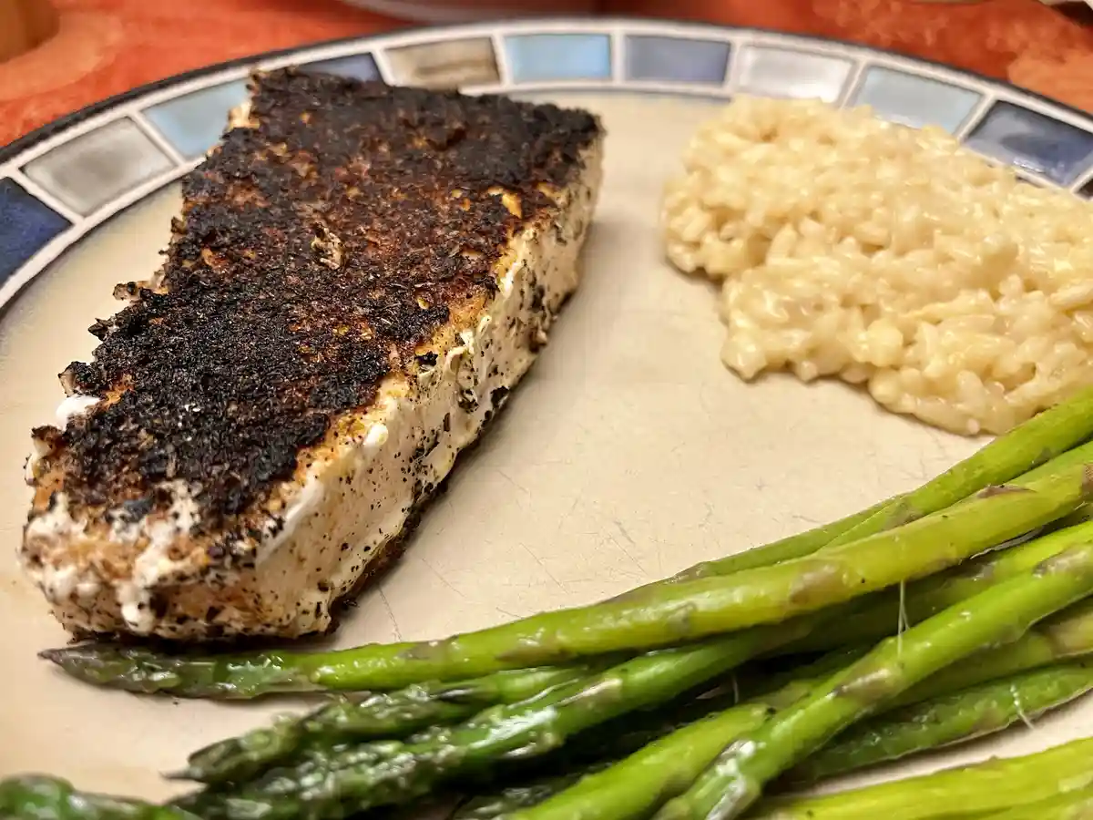 A close-up photo shows a plate with a heavily charred, blackened piece of salmon next to creamy risotto and green asparagus…