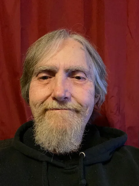 A close-up photo of an older man with gray hair and a full beard, wearing a black hoodie, smiling slightly while standing in…