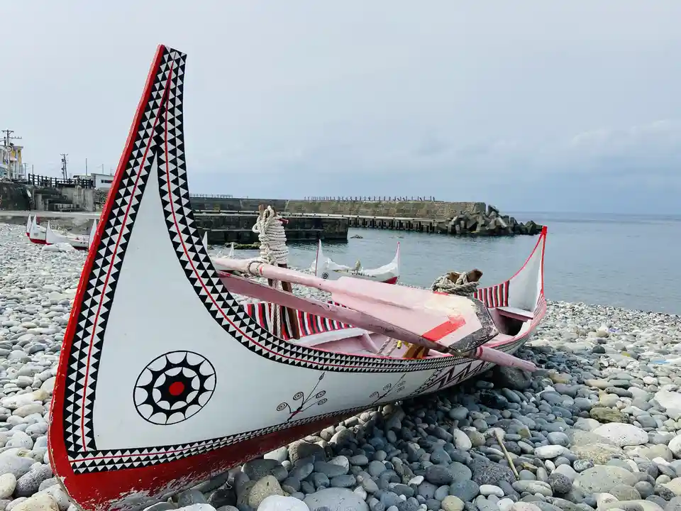 a traditional decorated canoe with red, white, and black geometric patterns rests on a pebble beach near the water, with…