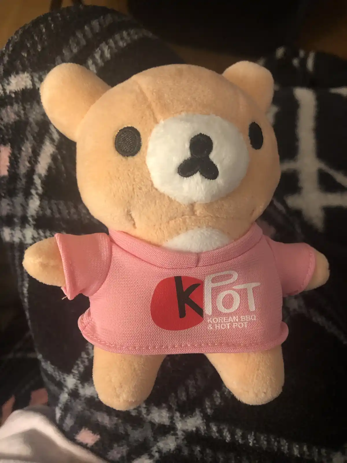A light brown plush teddy bear wearing a pink t-shirt with a logo that reads 'kpot korean bbq & hot pot' is sitting on a…