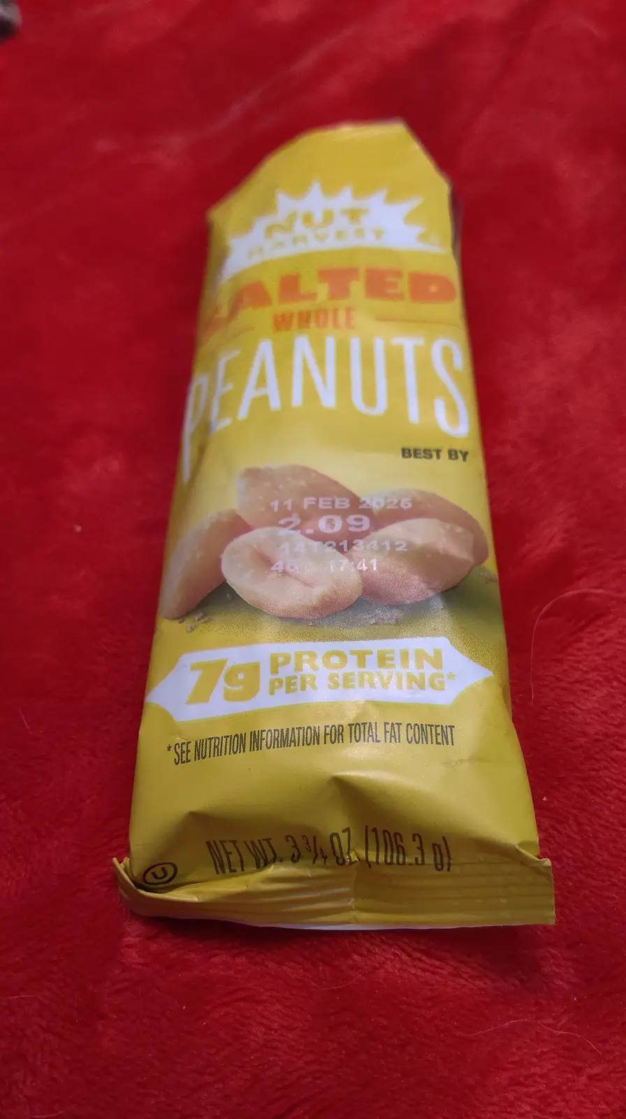 A yellow package of Nut Harvest Salted Whole Peanuts sits on a red textured surface. The 'best by' date is printed as '11…