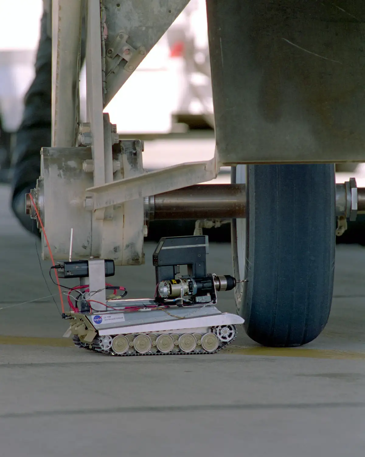 A small, tracked NASA robot labeled 'Tire Assault Vehicle' is shown positioned next to a large aircraft tire, with a drill…