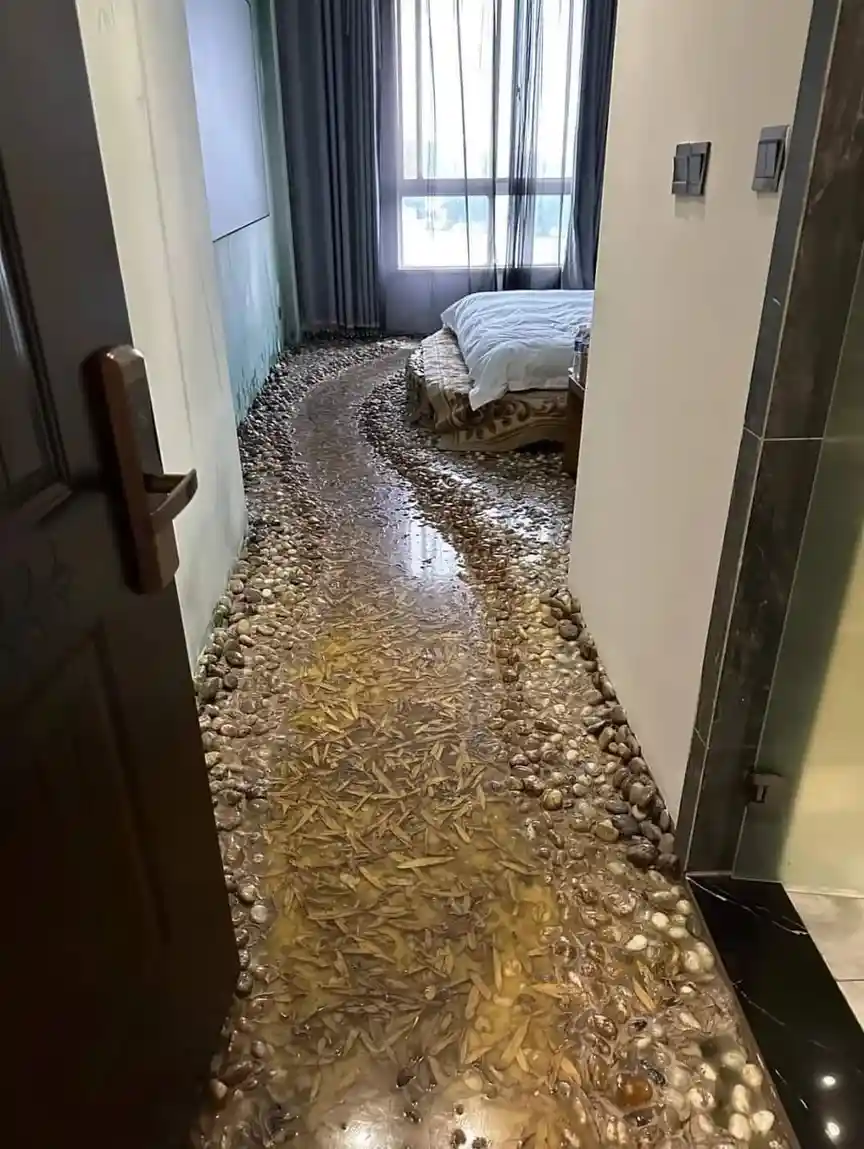 A hotel room hallway features a floor designed to look like a shallow river with pebbles and water, but it's filled with…