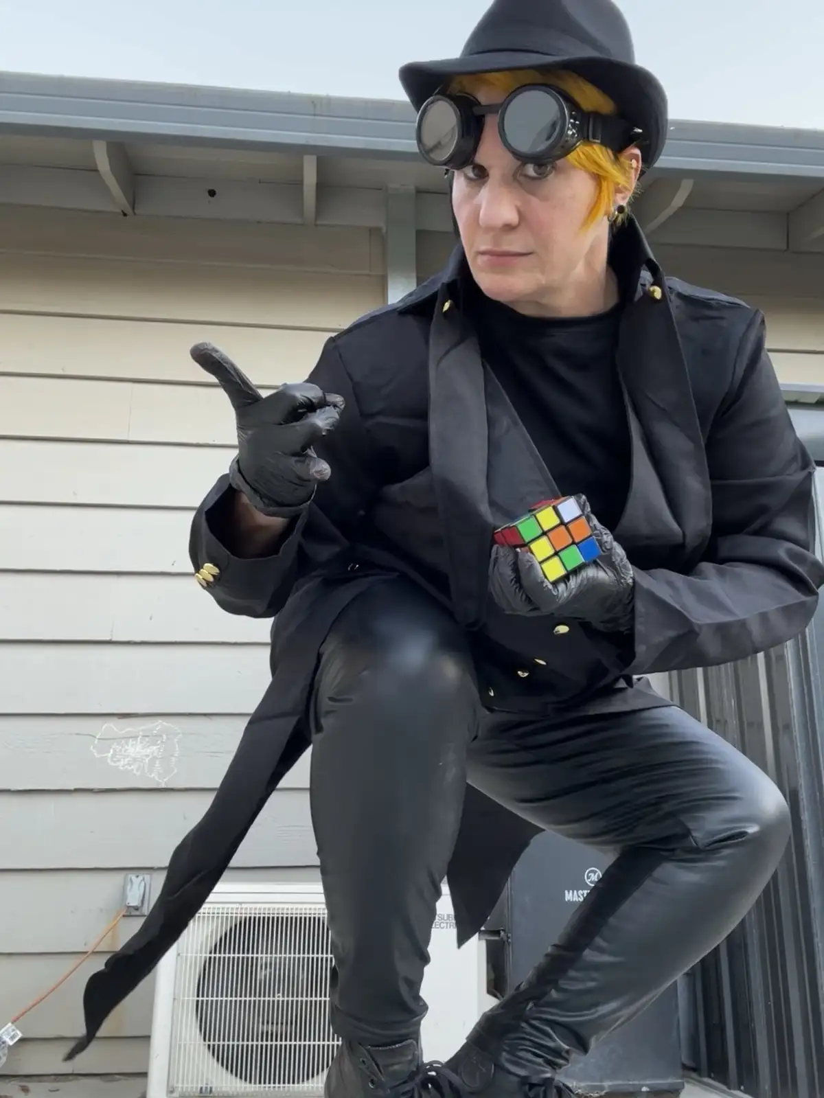 A person dressed in a black trench coat, leather pants, black gloves, and a black fedora with goggles, crouches while…