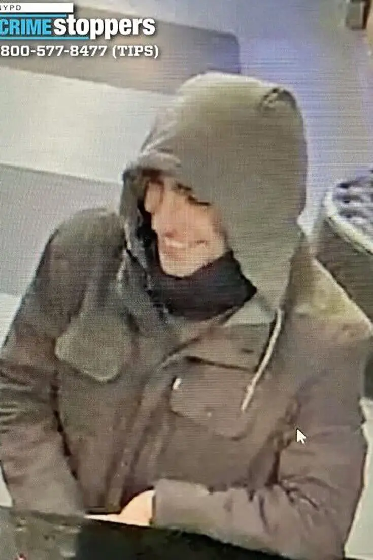 A grainy security camera image shows a person wearing a dark hooded jacket and a dark scarf, smiling while looking down. The…