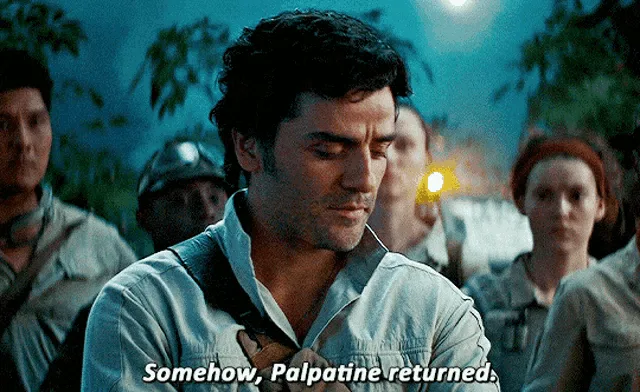 A man in a light blue shirt looks down with a resigned expression, surrounded by other people in a dimly lit outdoor…
