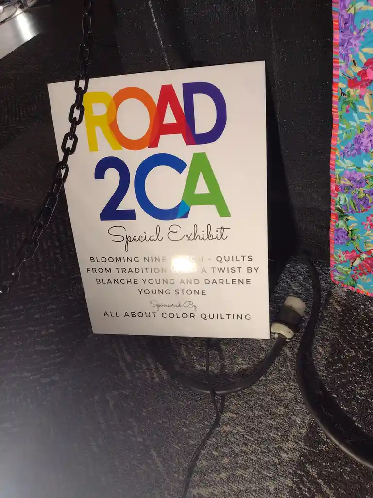 A white sign hanging by a black chain displays colorful text reading 'ROAD 2CA Special Exhibit' and details about quilts by…