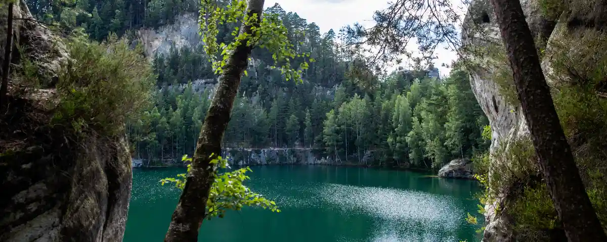 A scenic view of a calm, emerald-green lake surrounded by dense forests and rocky cliffs, framed by tree branches in the…