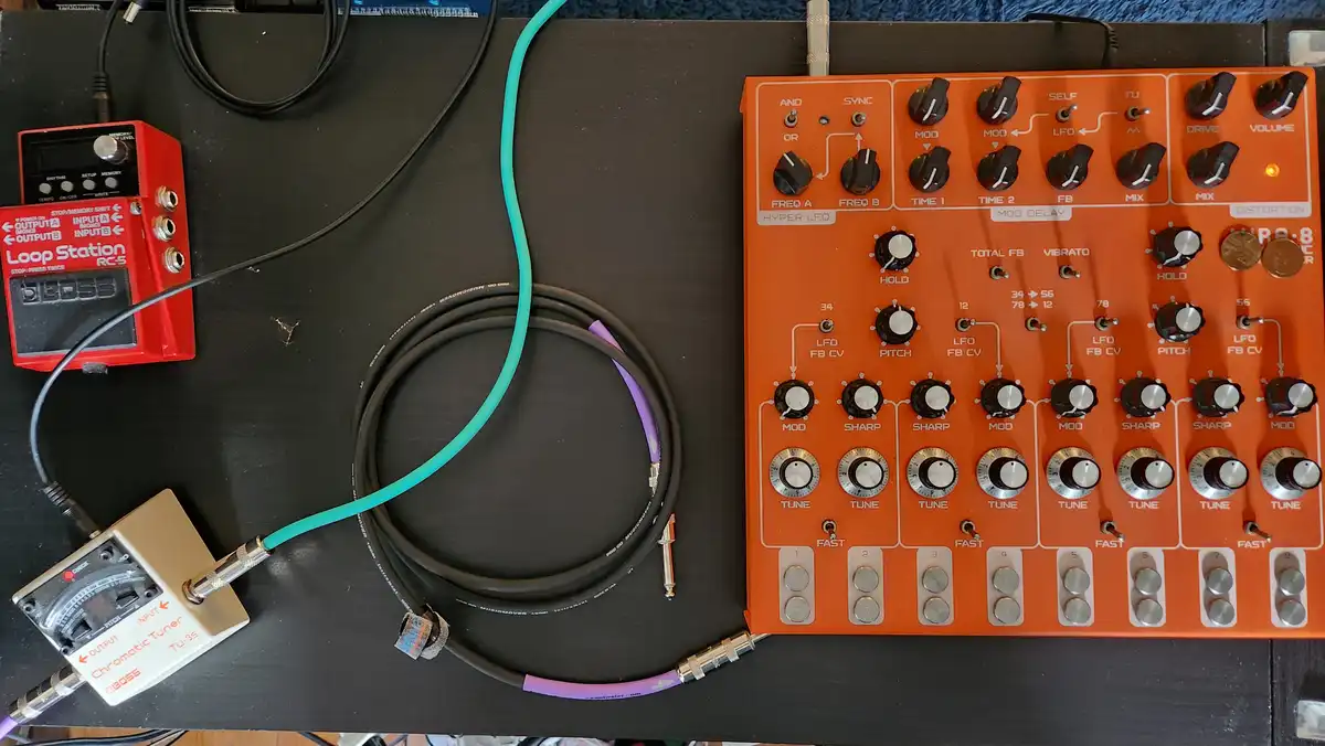 An overhead shot of a black surface with a red Boss Loop Station RC-5, a white Boss Chromatic Tuner TU-3S, and a large…