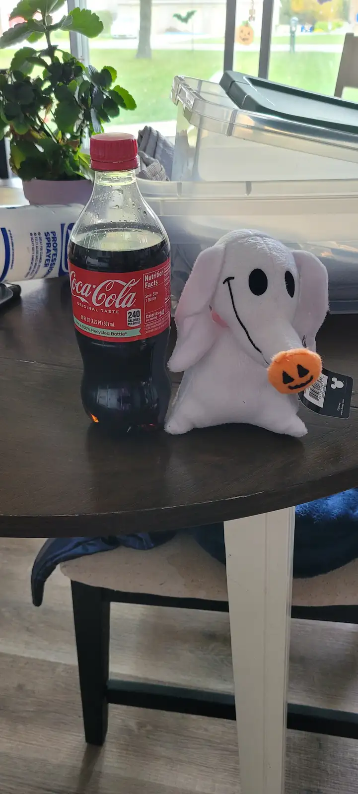 A partially consumed Coca-Cola bottle sits next to a white plush ghost toy with a jack-o'-lantern nose on a wooden table…
