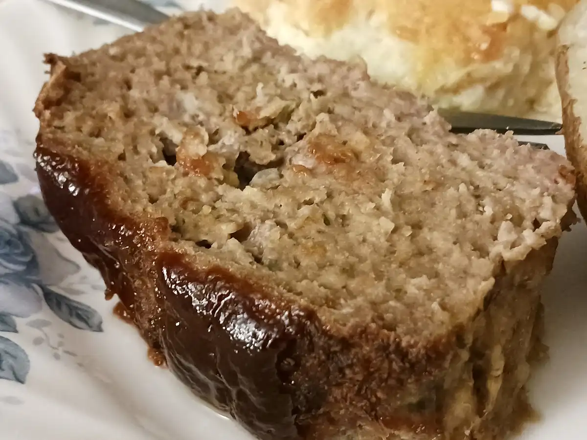 A close-up photo shows a slice of meatloaf with a glossy brown glaze on its crust, resting on a white plate with blue floral…