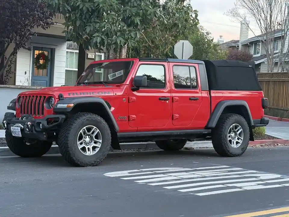 A red Jeep Rubicon pickup truck is parked on a residential street, positioned directly over a painted crosswalk. The vehicle…
