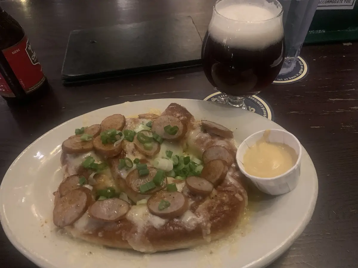 A close-up photo shows a round, cheesy pretzel-like bread topped with numerous slices of sausage and chopped green onions…