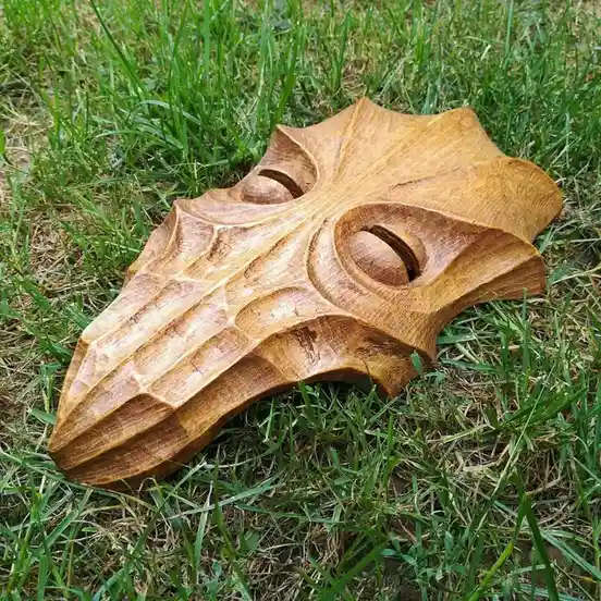 a hand-carved wooden plague doctor mask with a long beak and eye holes rests on green grass. The mask has intricate wood…