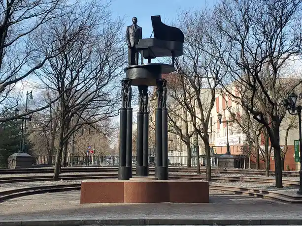 A bronze statue of a man in a suit standing beside a grand piano, mounted atop a pedestal supported by four pillars with…