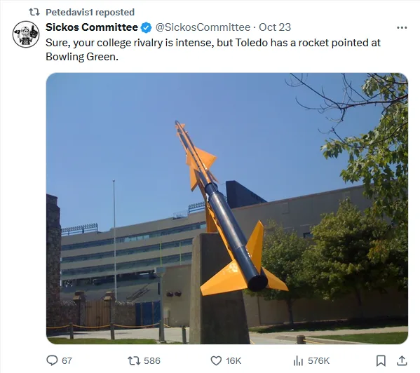 A large, orange and black rocket sculpture is mounted on a concrete pedestal outdoors, angled sharply upward and to the…