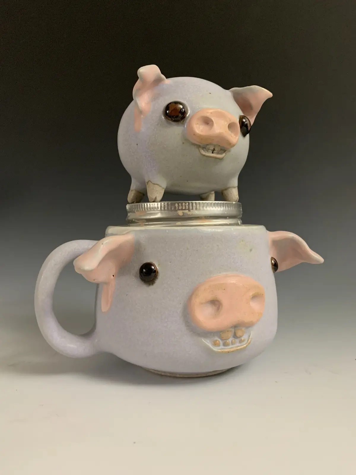 A ceramic mug shaped like a pig with pink ears, snout, and eyes, has a smaller pig figurine perched on its metal lid. The…