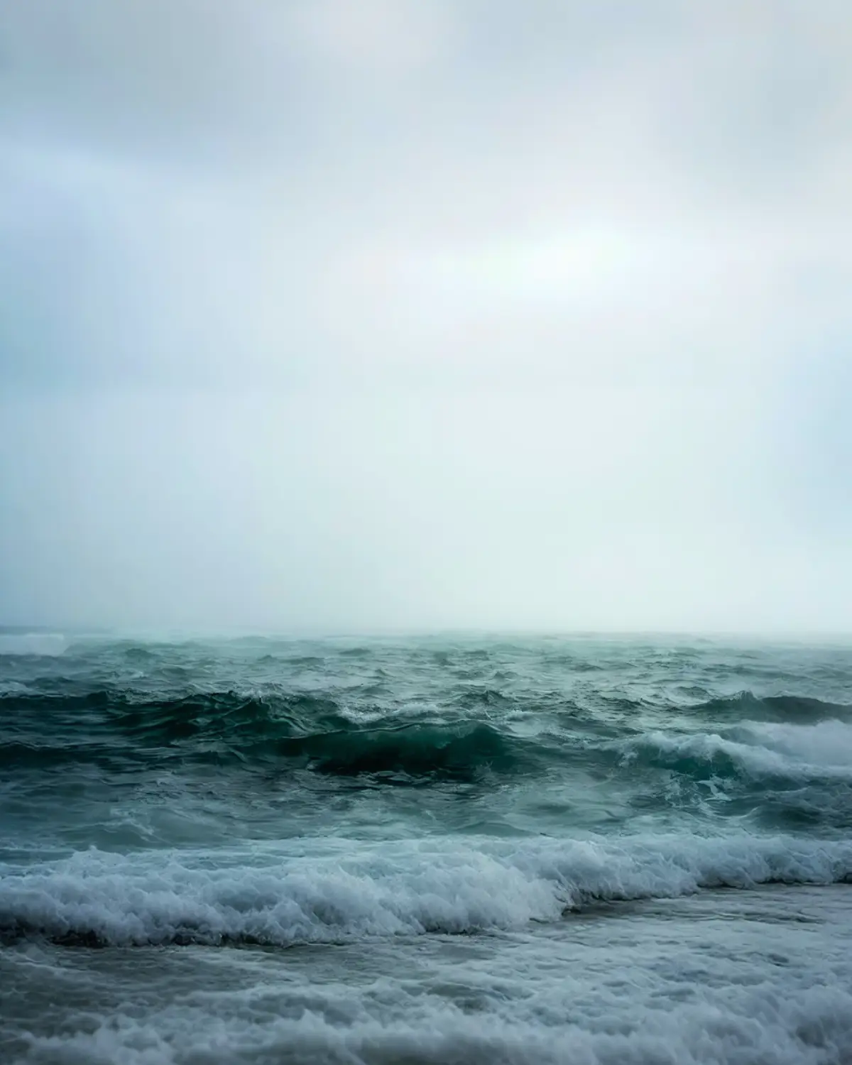 A moody, overcast seascape with turbulent, dark teal waves crashing onto a shore under a gray sky. The overlaid text reads…