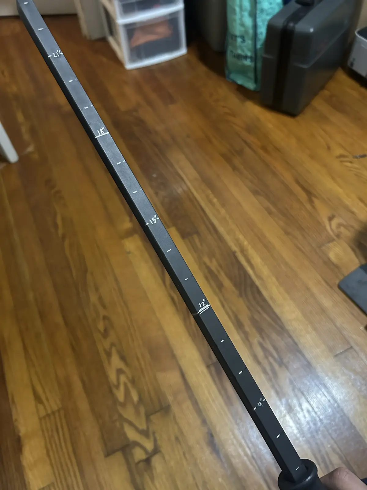 A close-up photo shows a black barbell lying diagonally across a wooden floor, with white inch markings visible along its…