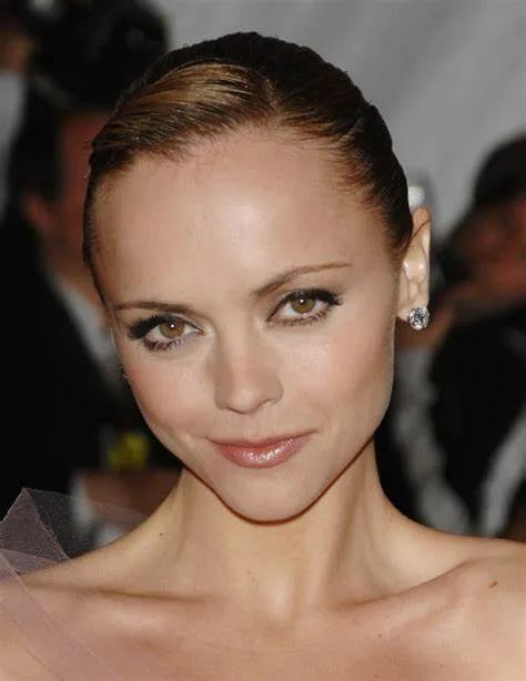 A close-up photo of a woman with slicked-back hair, heavy eyeliner, and a subtle smile, wearing diamond earrings and a sheer…