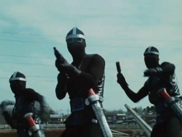 Three figures in black ninja-style outfits with white headbands and face coverings stand in a field, holding small handguns…