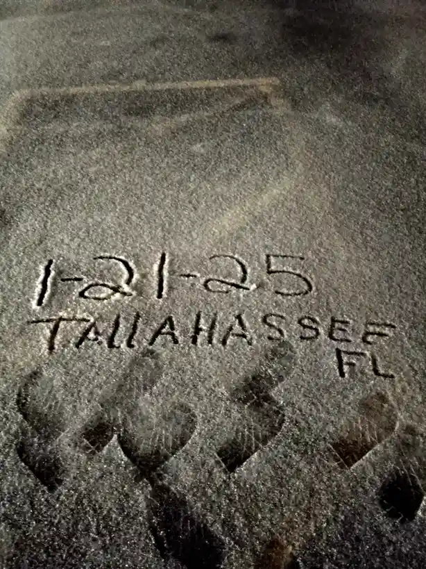 A close-up of a dark, textured surface with carved or etched text reading '1-21-25 TALLAHASSEE FL', with a faint outline of…