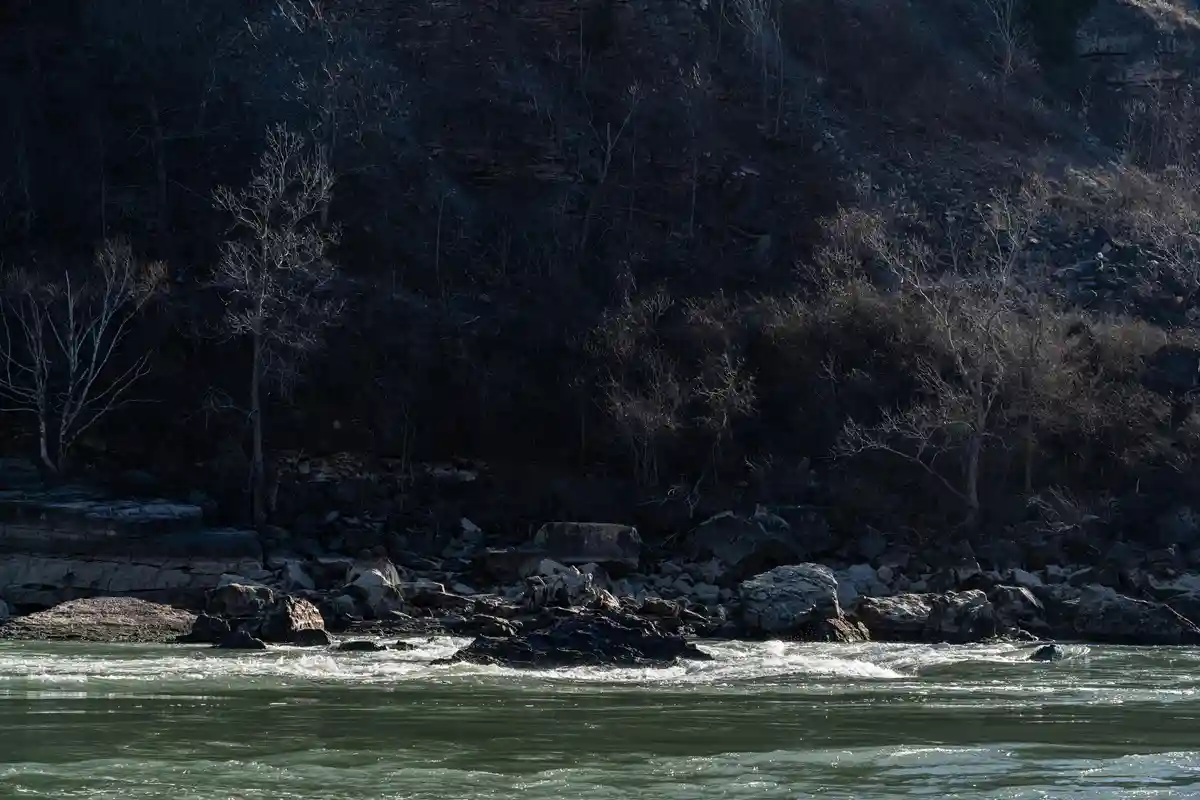 A dark, moody photo of a river flowing past rocky banks with bare winter trees on a steep, shadowed hillside. The overlaid…