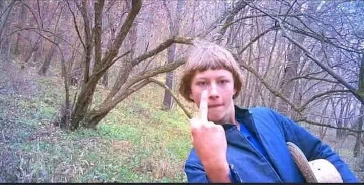 a person with short brown hair and a blue jacket stands in a wooded area with bare trees, pointing their index finger…