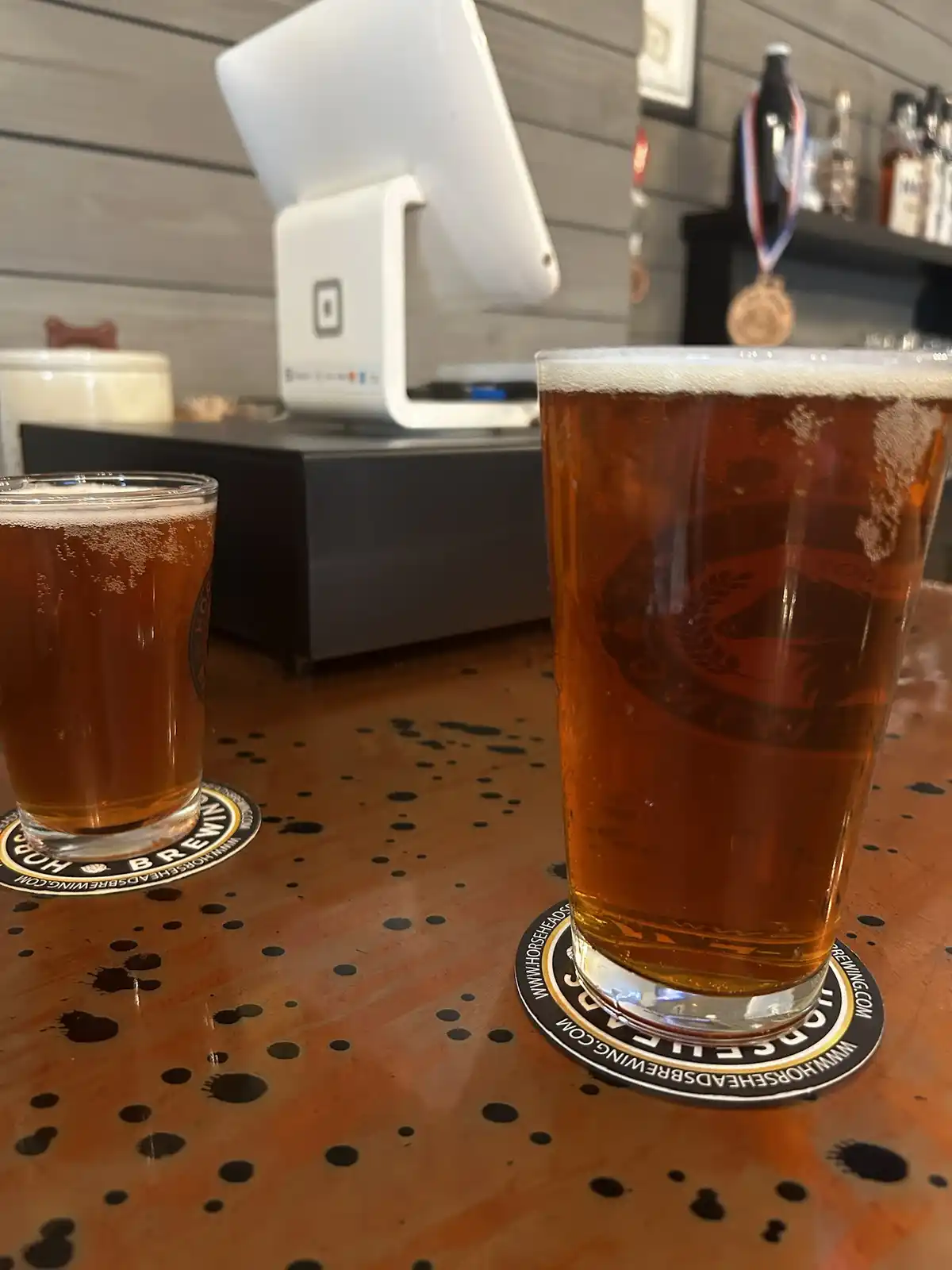 Two glasses of amber beer sit on branded coasters at a bar, one tall and one short, with a point-of-sale terminal and liquor…