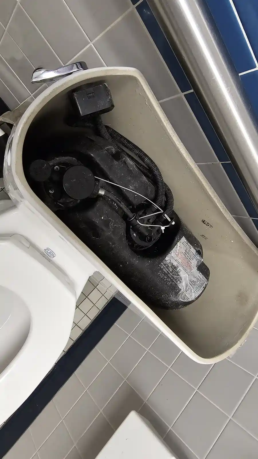 A close-up, angled view of a white toilet tank with its lid removed, revealing a large black pump and various wires and…