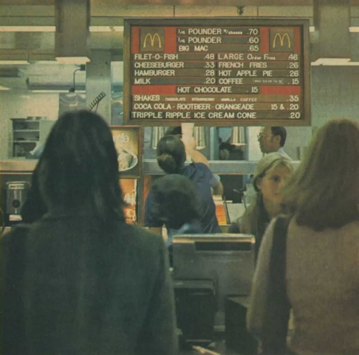 A vintage photo shows the back of customers at a 1970s McDonald's counter, looking at a menu board listing items like…